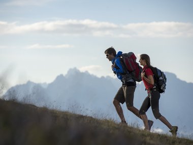 Two hikers with backpacks climbing a hill with mountains in background