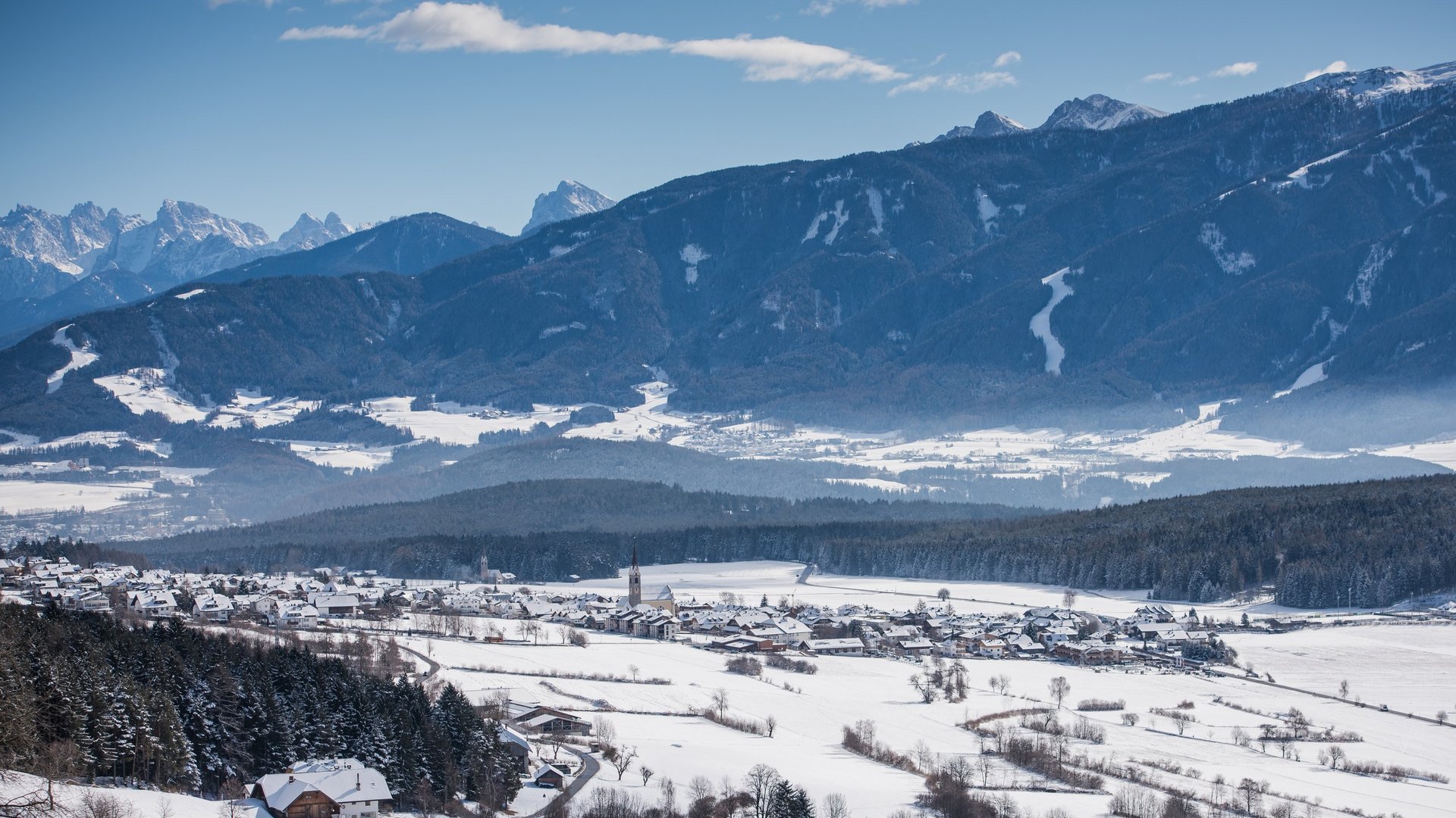 Schneebedecktes Dorf vor bewaldeten Hügeln und Bergen unter klarem Himmel